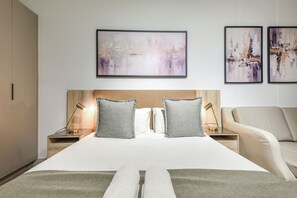 Une chambre d’hôtel avec un grand lit, deux tables de chevet avec des lampes, un canapé et trois tableaux abstraux encadrés accrochés au mur.