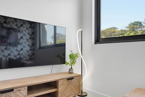 A modern living room with a flat-screen TV mounted on the wall, a wooden entertainment unit, a floor lamp, and a window with a view of trees. - Zimbali Lakes Studio | Rooftop Pool, Balcony (Westbrook)