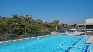 En klar blå swimmingpool med glasafskærmning, omgivet af frodigt grønt og en moderne bygning.