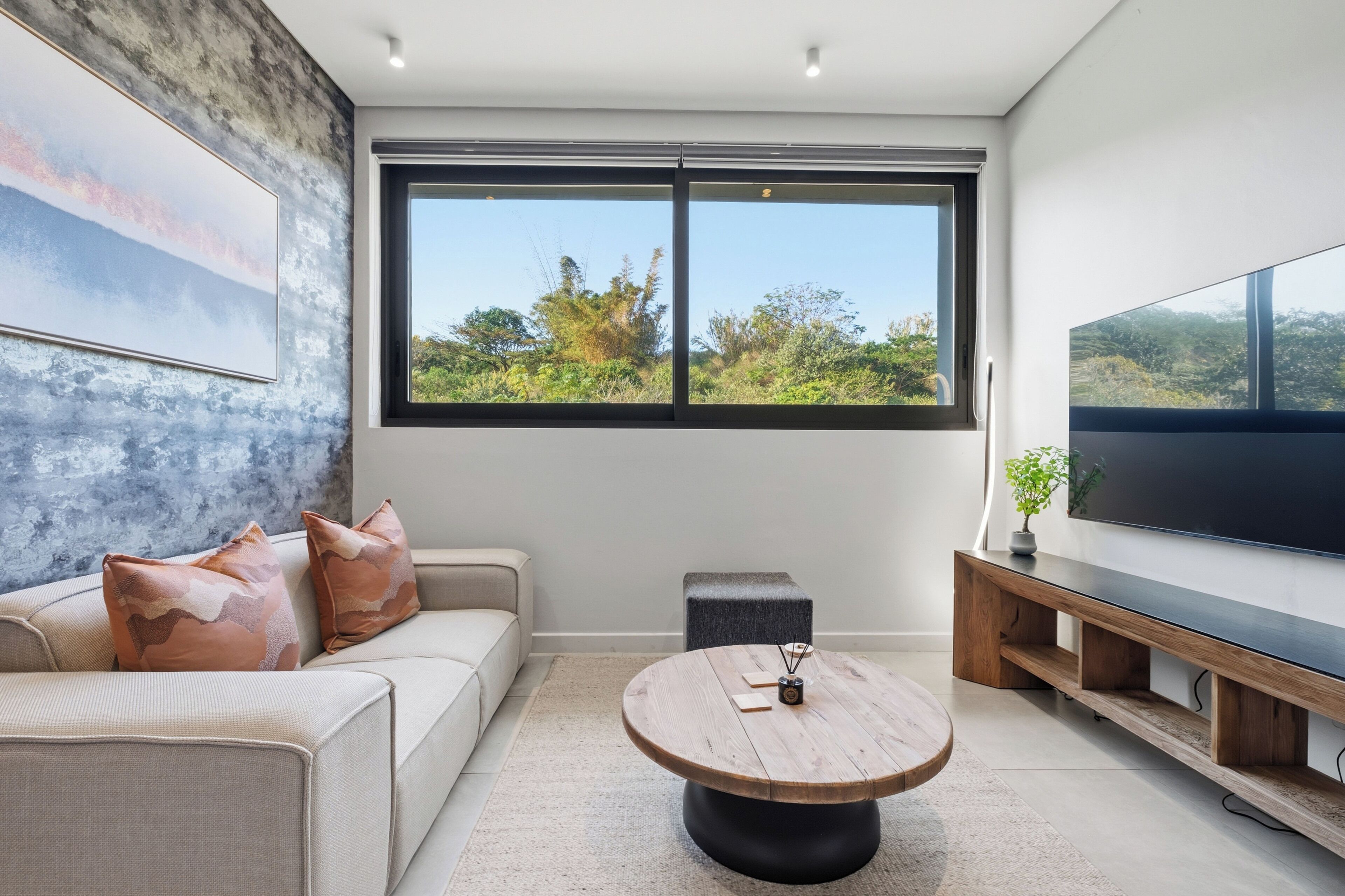 A modern living room with a large window, a flat-screen TV, a round wooden coffee table, and a sofa with patterned cushions.
