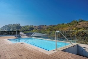 Ein Schwimmbecken mit Holzterrasse, umgeben von einem Glaszaun, mit Blick auf eine Hügellandschaft mit Häusern.