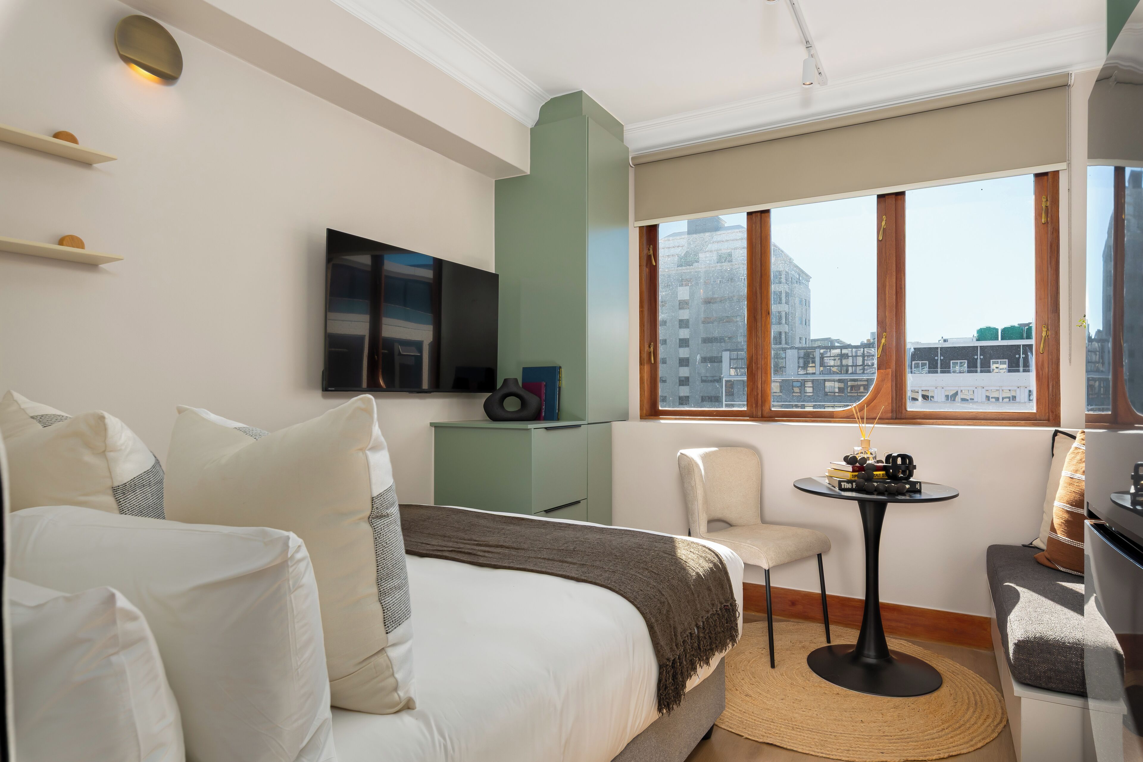 A hotel room with a bed, a television, a small table with books, a chair, and a window with a city view.