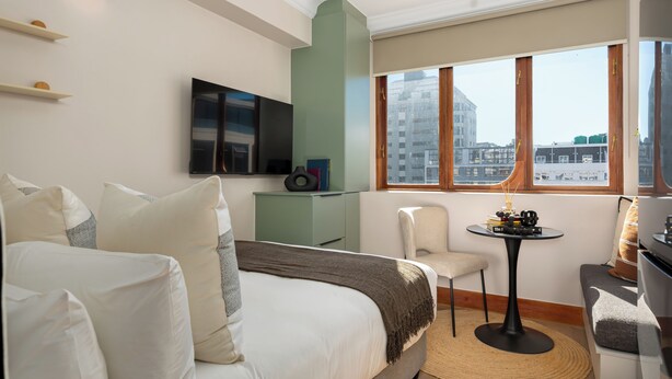 Ein Hotelzimmer mit einem Bett, einem Fernseher, einem kleinen Tisch mit Büchern, einem Sessel und einem Fenster mit Blick auf die Stadt.