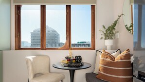 Ein stilvoller Wohnraum mit Blick auf eine Stadtlandschaft durch ein großes Fenster, ein runder Tisch mit Büchern und einer Kerze, ein bequemer Sessel und eine Couch mit gemusterten Kissen.