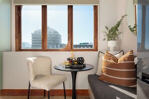 Ein stilvoller Wohnraum mit Blick auf eine Stadtlandschaft durch ein großes Fenster, ein runder Tisch mit Büchern und einer Kerze, ein bequemer Sessel und eine Couch mit gemusterten Kissen.