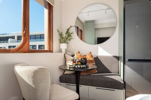 Ein modernes Zimmer mit einem runden Spiegel, einem kleinen Tisch mit Büchern und einer Pflanze, einem Sofa und Blick auf ein Gebäude.