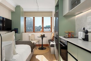 Ein kompaktes Hotelzimmer mit Küchenzeile, einer Sitzecke mit kleinem Tisch, einem Sofa und einem großen Fenster mit Blick auf die Stadt.