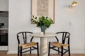 A dining area with a round marble table, two black chairs with woven seats, a vase with green plants, and a wall-mounted light fixture. - Dolce Vita, Stylish Sea Point Retreat (Cape Town)
