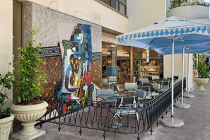 Un área de estar al aire libre con mesas y sillas, un mural grande en la pared y una planta decorativa.