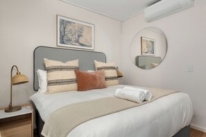 Cama bem arrumada com edredom branco e várias almofadas, abajur, ar condicionado fixado na parede e uma pintura de paisagem emoldurada sobre a cama.