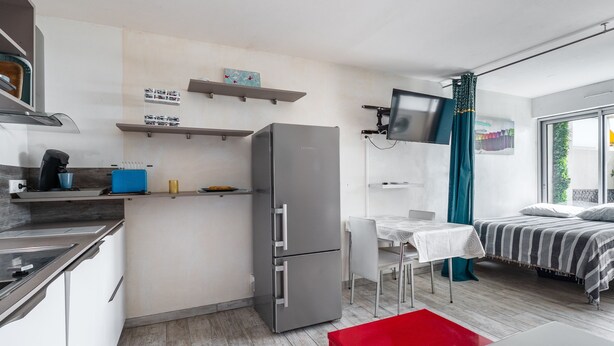 A compact kitchen with a refrigerator, a coffee machine, and a microwave. There is a dining area with a table and chairs, and a bed with a striped blanket. - Le Jardin de La Plage - Climatisé et plage à 100 m (Canet-en-Roussillon)
