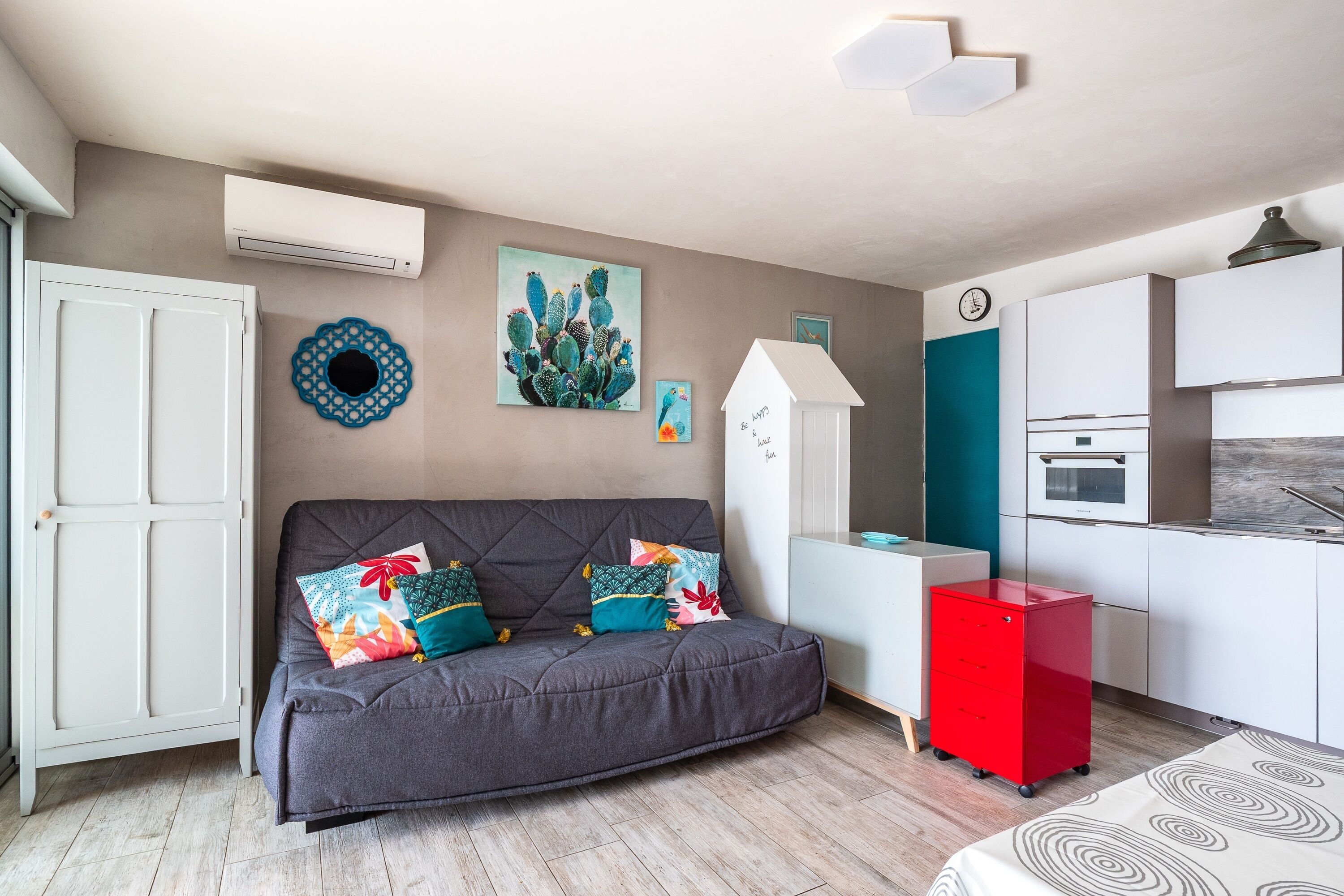 Un espace de vie compact comprenant un canapé, un petit coin cuisine et un tableau décoratif représentant un cactus.