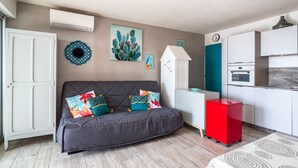 A compact living space with a sofa, a small kitchen area, and a decorative cactus painting. - Le Jardin de La Plage - Climatisé et plage à 100 m (Canet-en-Roussillon)