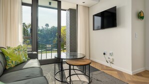 Ein modernes Wohnzimmer mit einer Couch, einem Glastisch, einem an der Wand befestigten Fernseher und Blick ins Grüne.