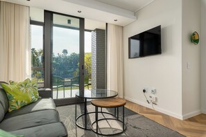 Ein modernes Wohnzimmer mit einer Couch, einem Glastisch, einem an der Wand befestigten Fernseher und Blick ins Grüne.