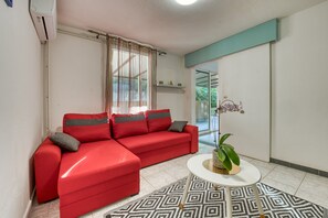Un canapé rouge avec des coussins gris, une table basse blanche et un tapis à motifs dans une pièce aux murs blancs et dotée d’une grande fenêtre.
