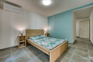 A bedroom with a wooden bed, two bedside tables with lamps, a wall-mounted air conditioner, and a tiled floor. - La Case Coco - Bananes - Appartement climatisé (Saint Joseph)