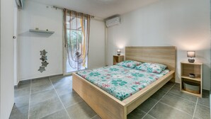 A bedroom with a wooden bed, a nightstand with a lamp, a wall-mounted air conditioner, and a window with curtains. - La Case Coco - Bananes - Appartement climatisé (Saint Joseph)