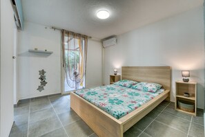 A bedroom with a wooden bed, a nightstand with a lamp, a wall-mounted air conditioner, and a window with curtains. - La Case Coco - Bananes - Appartement climatisé (Saint Joseph)