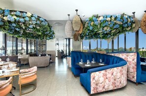 A modern dining area with blue upholstered seating, a long table with wine glasses, and decorative hanging plants. - Effortless Luxury in Rosebank (Johannesburg)
