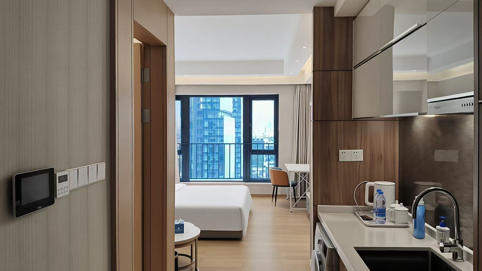 A modern hotel room with a bed, a small table, a kitchenette, and a view of the city.