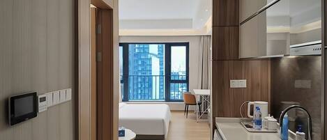 A modern hotel room with a bed, a small table, a kitchenette, and a view of the city.