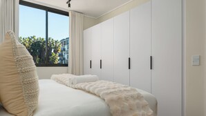 A bedroom with a large bed, a window with curtains, and a view of palm trees outside. - Park Place, Designer Retreat, Lake Views (Cape Town)