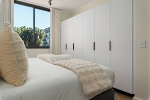 A bedroom with a large bed, a window with curtains, and a view of palm trees outside. - Park Place, Designer Retreat, Lake Views (Cape Town)