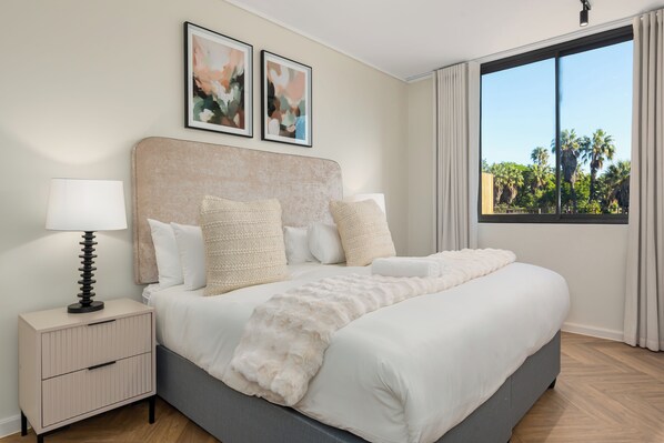 Une chambre à coucher avec un lit, une table de chevet, une lampe et une fenêtre donnant sur des palmiers.