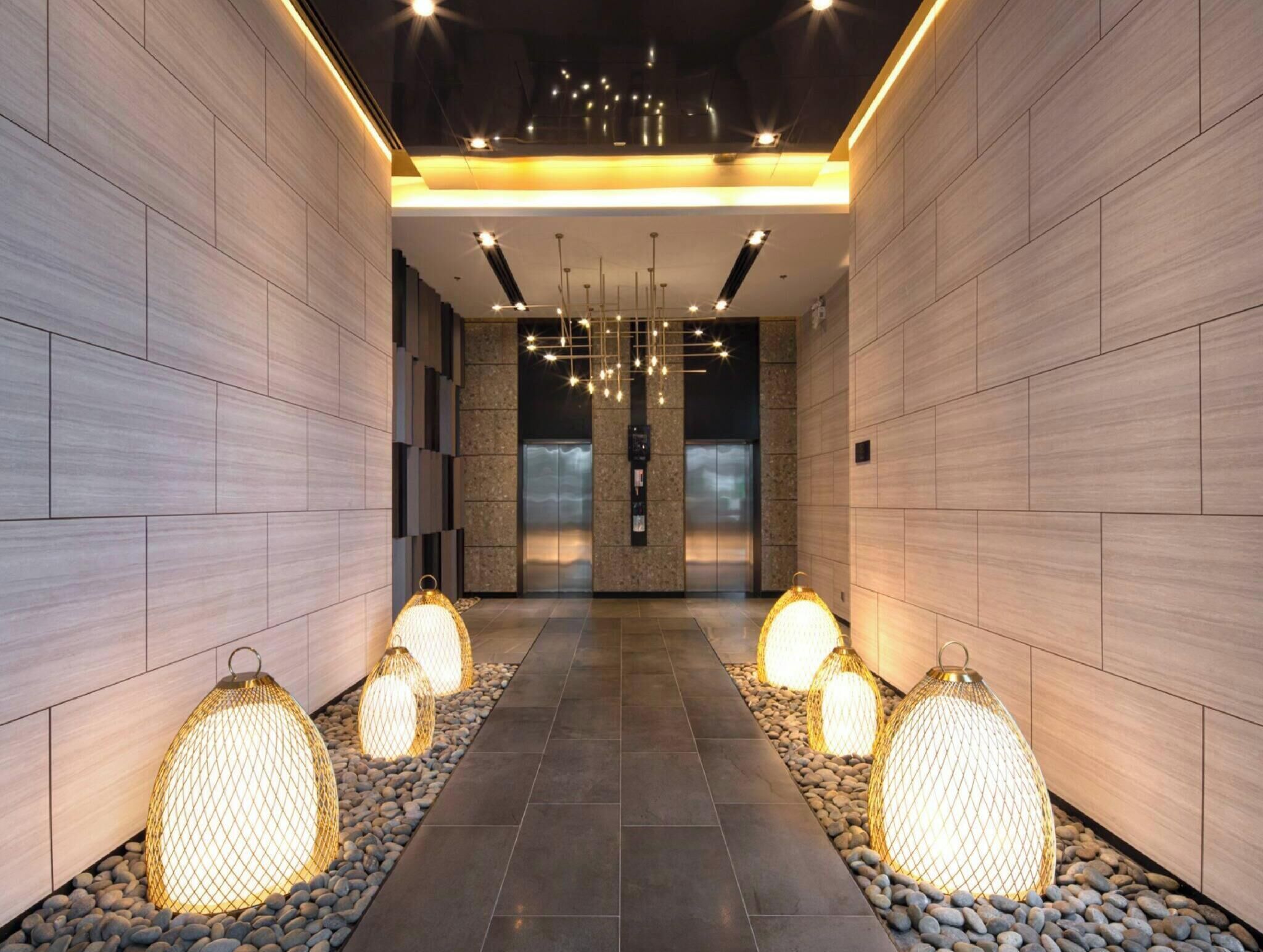 A modern hallway with a central elevator, decorative hanging lights, and a patterned floor.