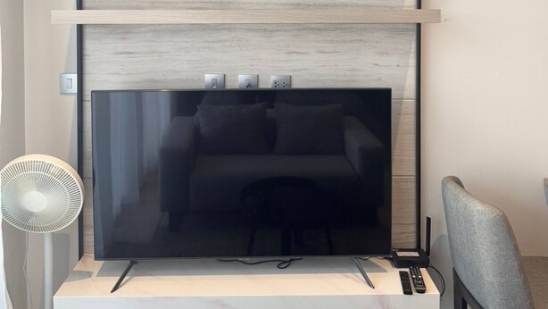 Ett modernt vardagsrum med en stor tv-skärm på ett vitt tv-bord, en grå fåtölj och ett litet bord med fjärrkontroller.