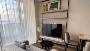 Ett modernt vardagsrum med en grå soffa, ett svart soffbord, en tv på en hylla i trä och en matplats med ett litet bord och stolar.