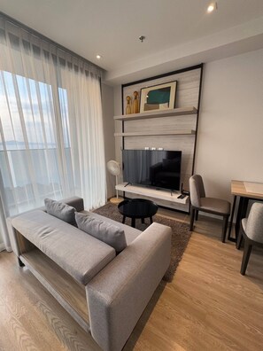 A modern living room with a grey sofa, a black coffee table, a TV on a wooden shelf, and a dining area with a small table and chairs. - Seaview Beach At A Great Price (Pattaya)