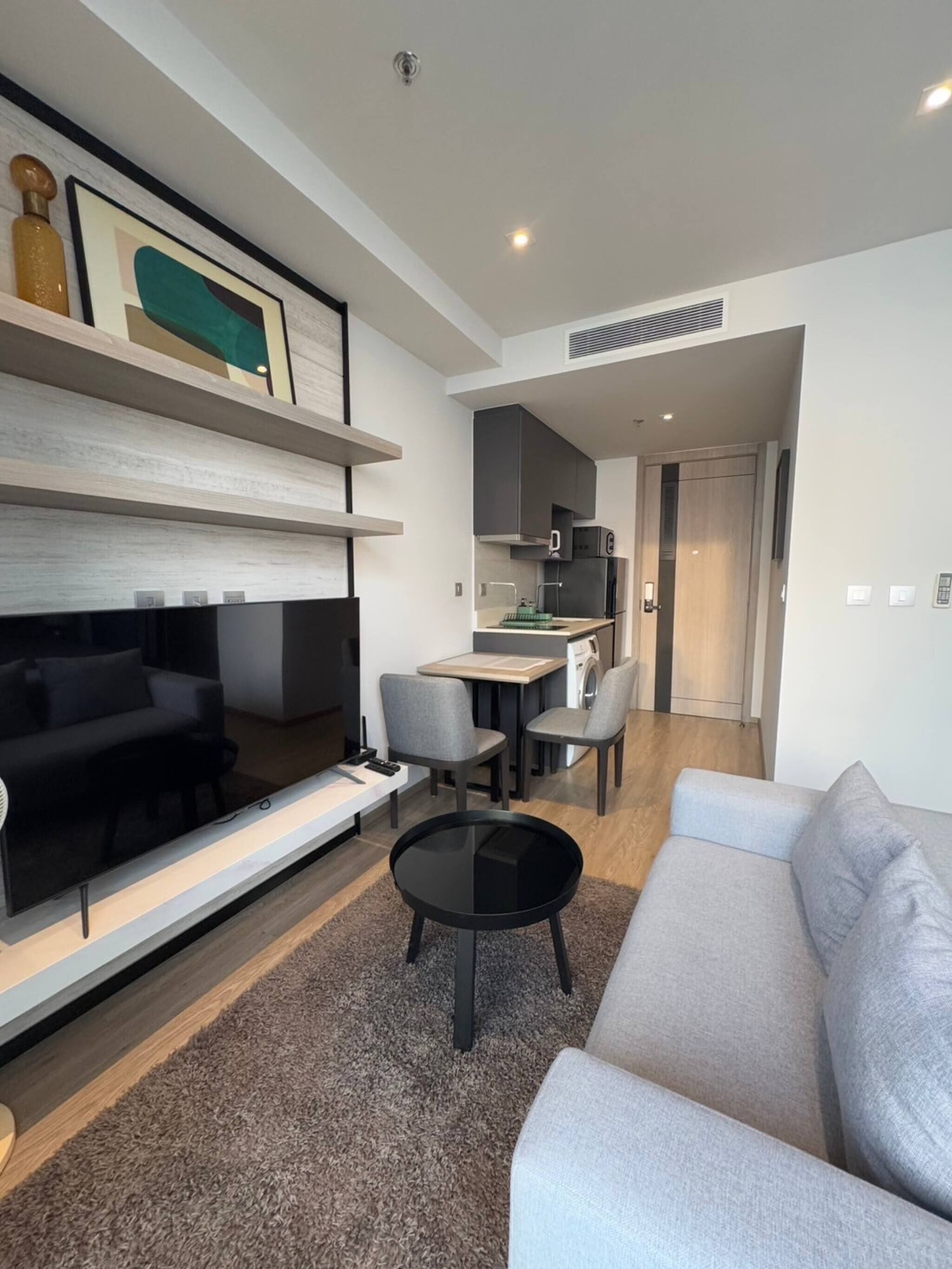 Una sala de estar moderna con un televisor de pantalla plana, un sofá gris, una mesa de centro y un comedor con sillas, con una cocina al fondo.