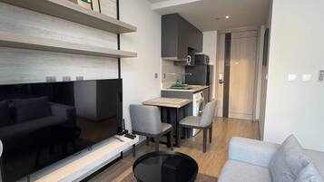 A modern living room with a flat-screen TV, a gray sofa, a coffee table, and a dining area with chairs and a kitchen in the background.