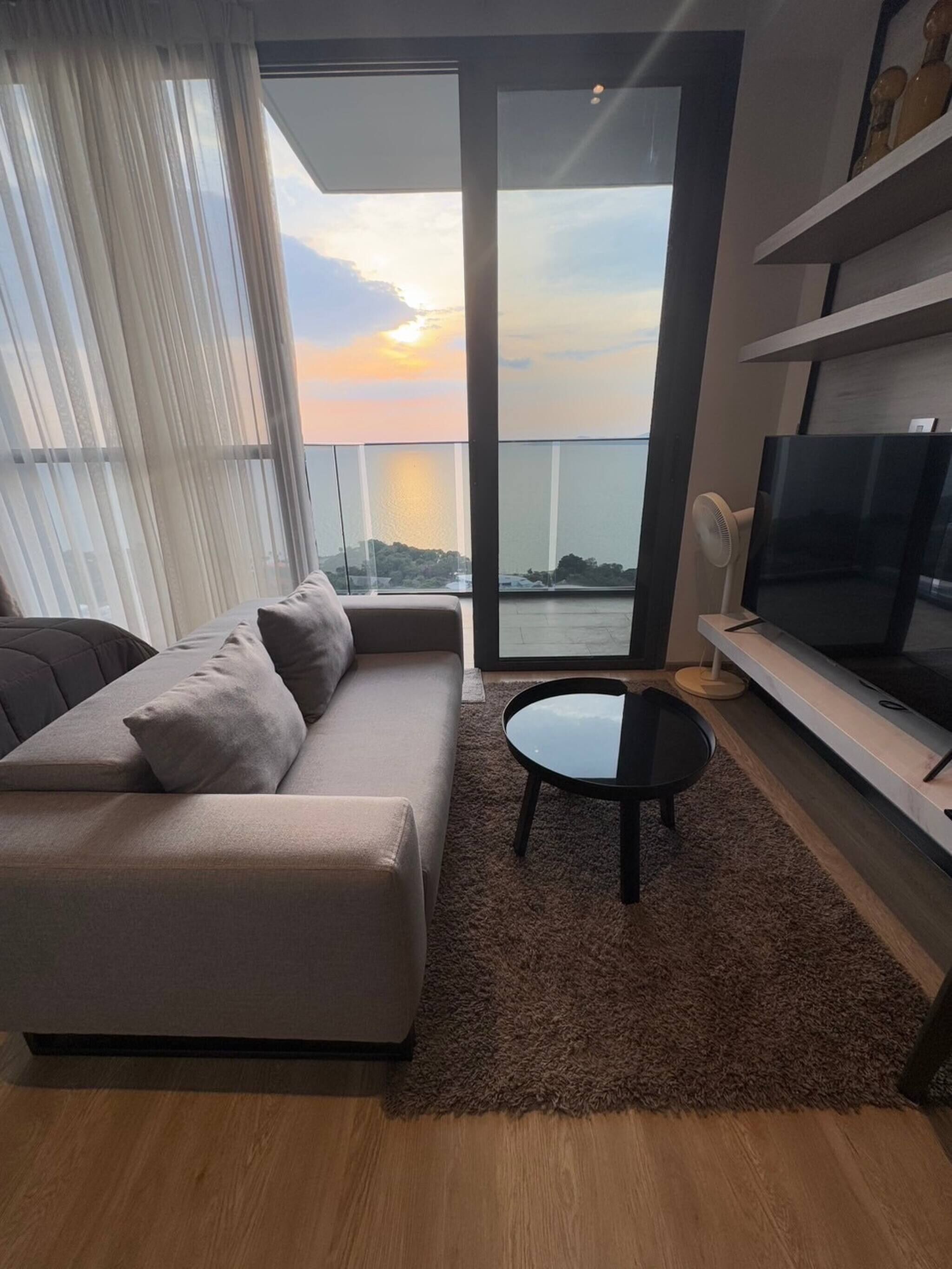 A modern living room with a sofa, a coffee table, and a TV. It features a large window with a view of the city skyline and a balcony.