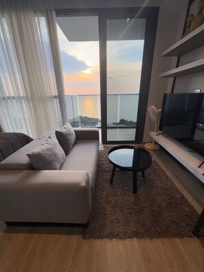 A modern living room with a sofa, a coffee table, and a TV. It features a large window with a view of the city skyline and a balcony. - Seaview Beach At A Great Price (Pattaya)