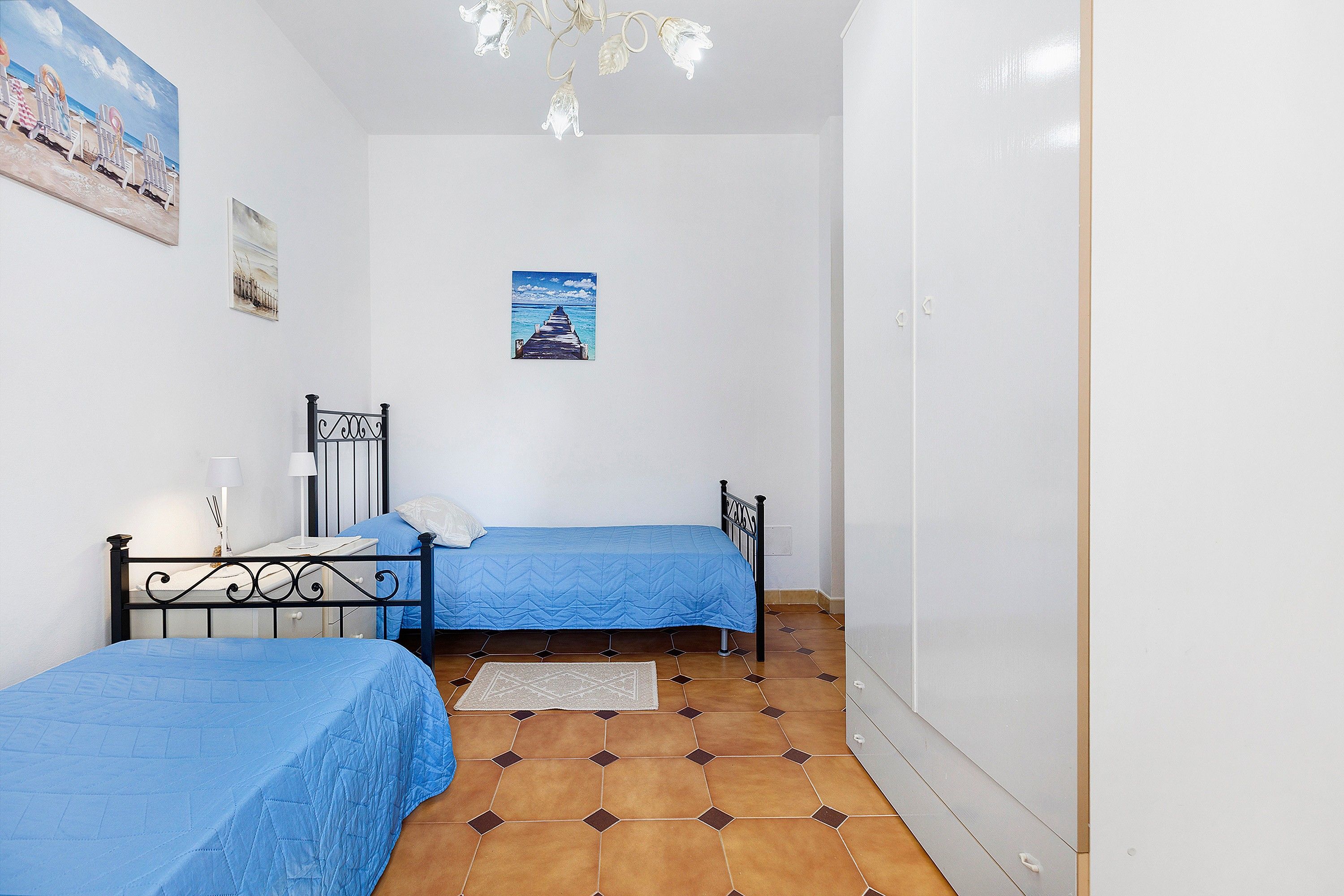 A bedroom with two beds, a small table, a nightstand, a poster on the wall, and a tiled floor.