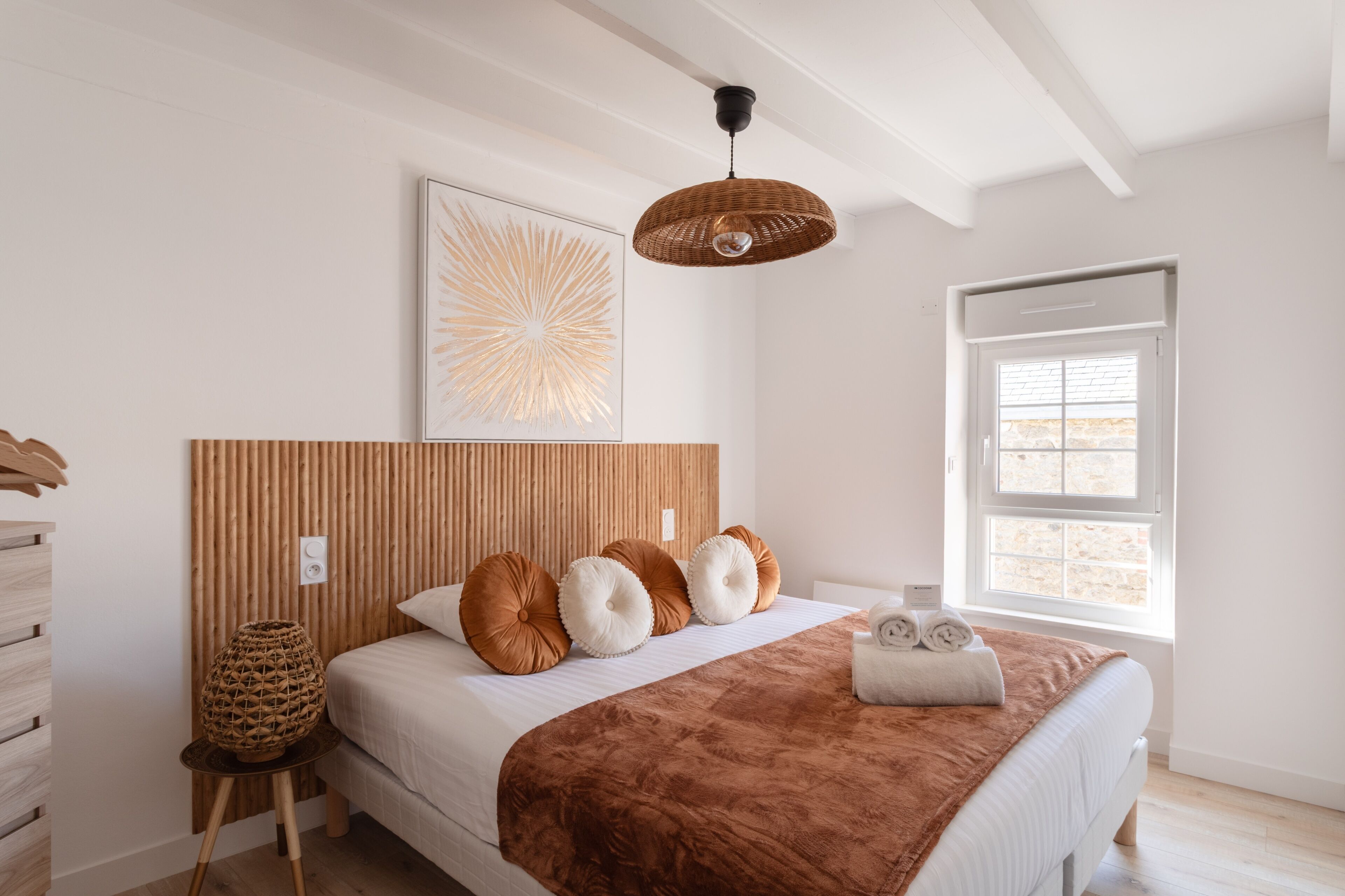Une chambre avec une tête de lit en lattes de bois, une grande fenêtre et un luminaire en matière tressée.