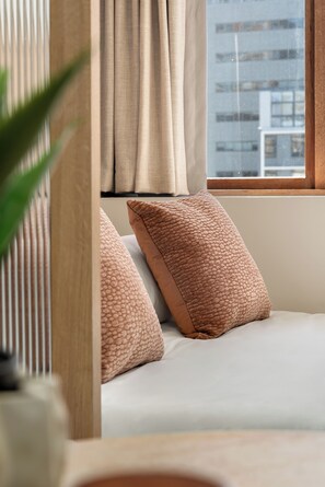 Une chambre à coucher avec un lit, deux oreillers, une fenêtre avec des rideaux et une vue sur les bâtiments à l’extérieur.