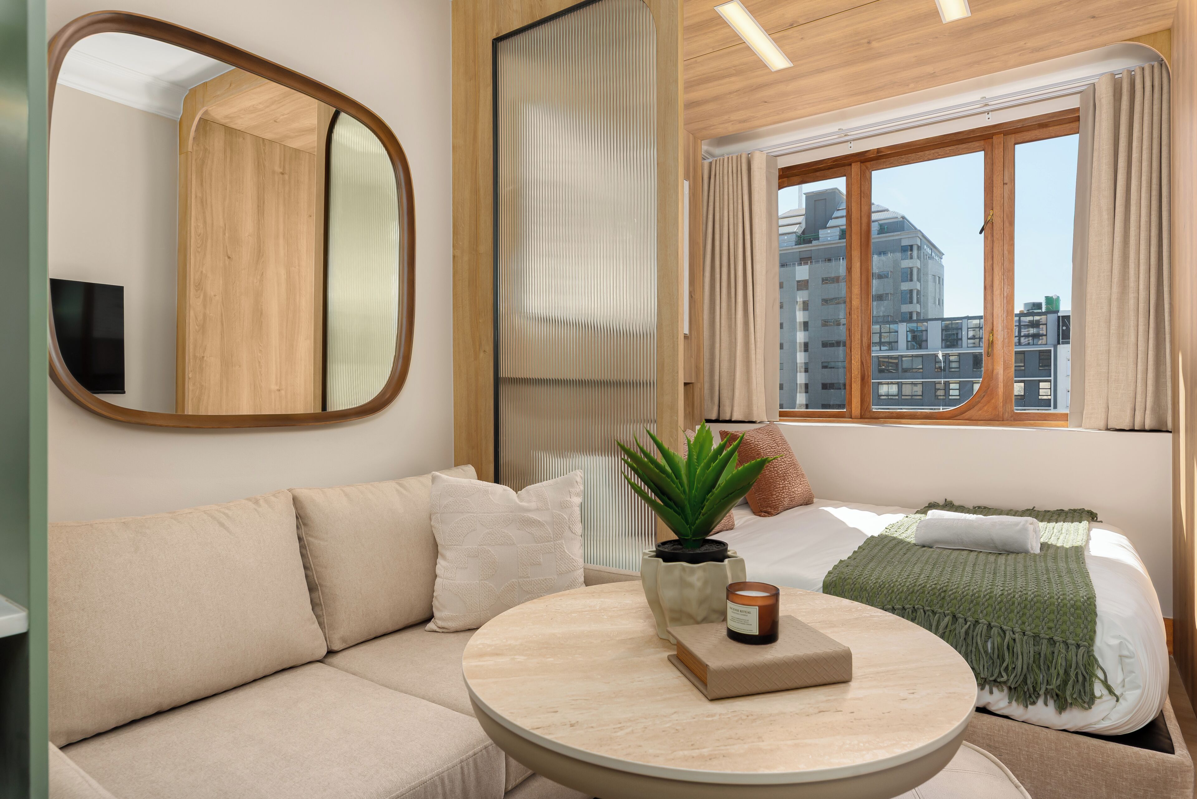 A modern hotel room with a sofa, a round table, a potted plant, a candle, and a view of the city through a window.
