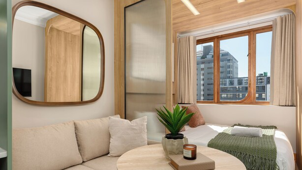 Ein modernes Hotelzimmer mit einer Sitzecke, einem runden Tisch, einer Zimmerpflanze, einer Kerze und Blick auf die Stadt durch ein Fenster.