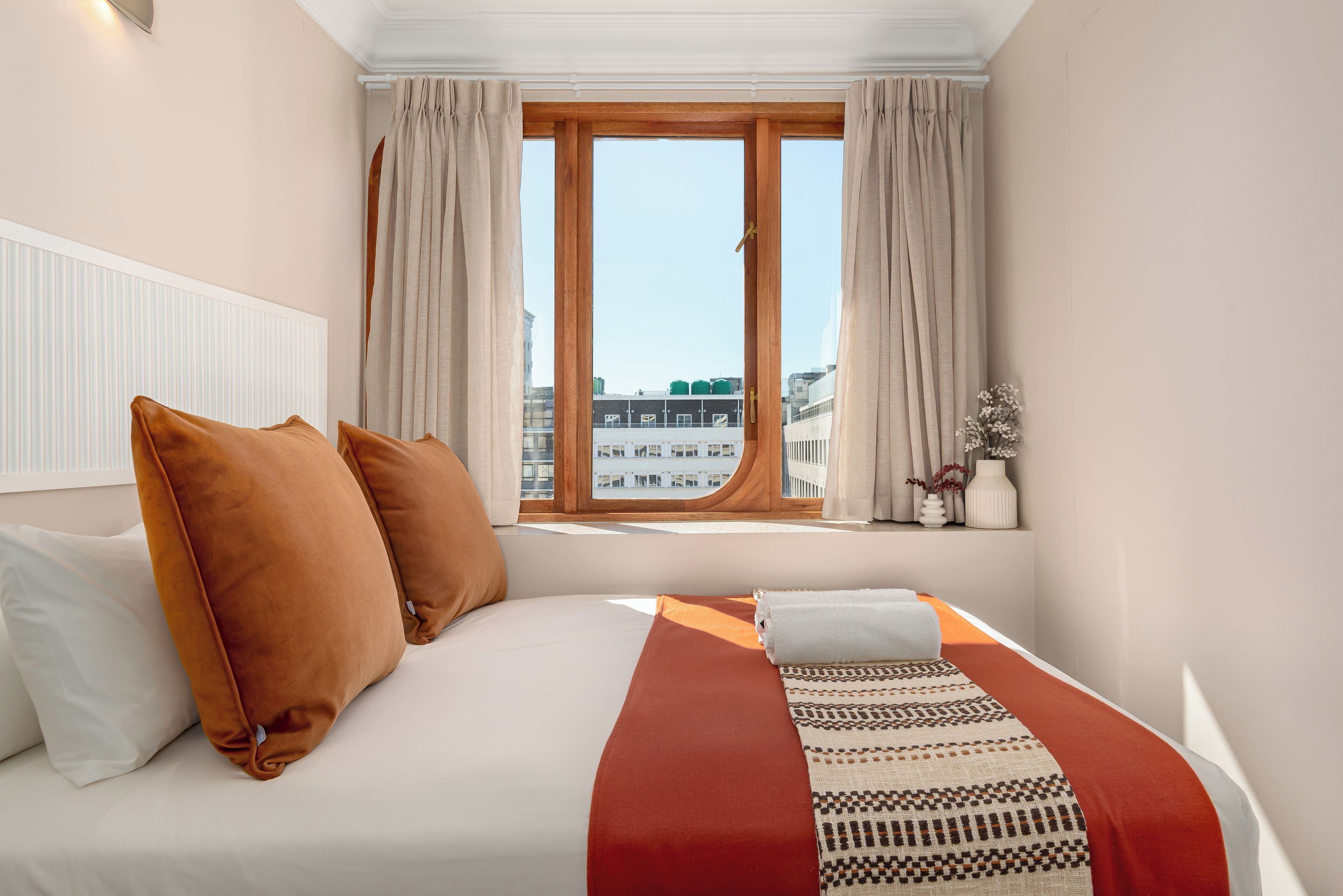 A hotel room with a large bed, a window with curtains, a radiator, and a view of buildings outside.