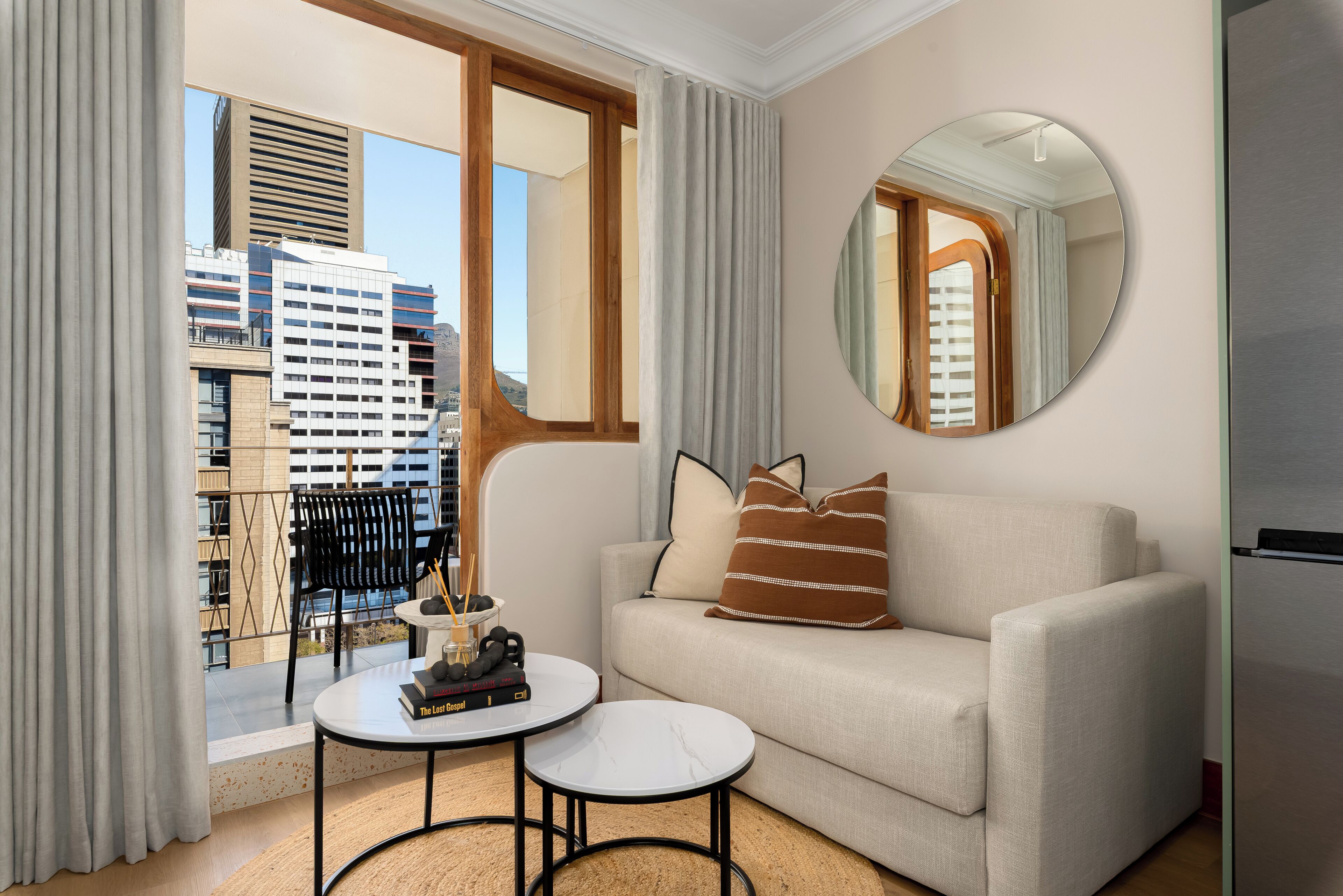 Ein modernes Wohnzimmer mit einer Couch, einem runden Spiegel, einem kleinen Tisch mit Büchern und Blick auf die Stadtlandschaft durch große Fenster.
