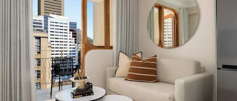 A modern living room with a sofa, a round mirror, a small table with books, and a view of the cityscape through large windows.