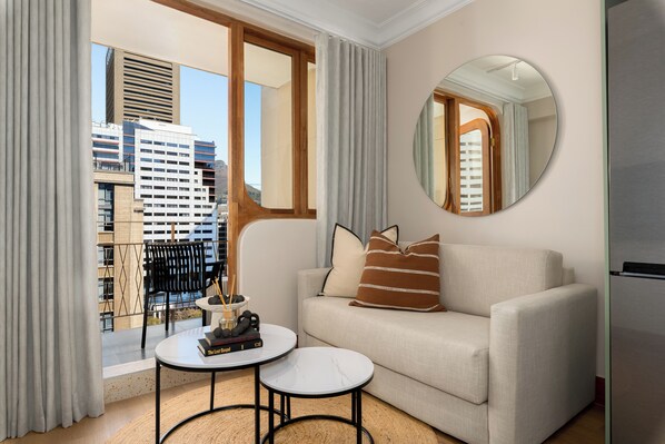 Una sala de estar moderna con un sofá, un espejo redondo, una mesita con libros y vistas a la ciudad a través de amplias ventanas.