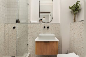 Un baño moderno con ducha independiente, un lavamanos blanco con gabinete de madera y un espejo sobre el lavamanos.