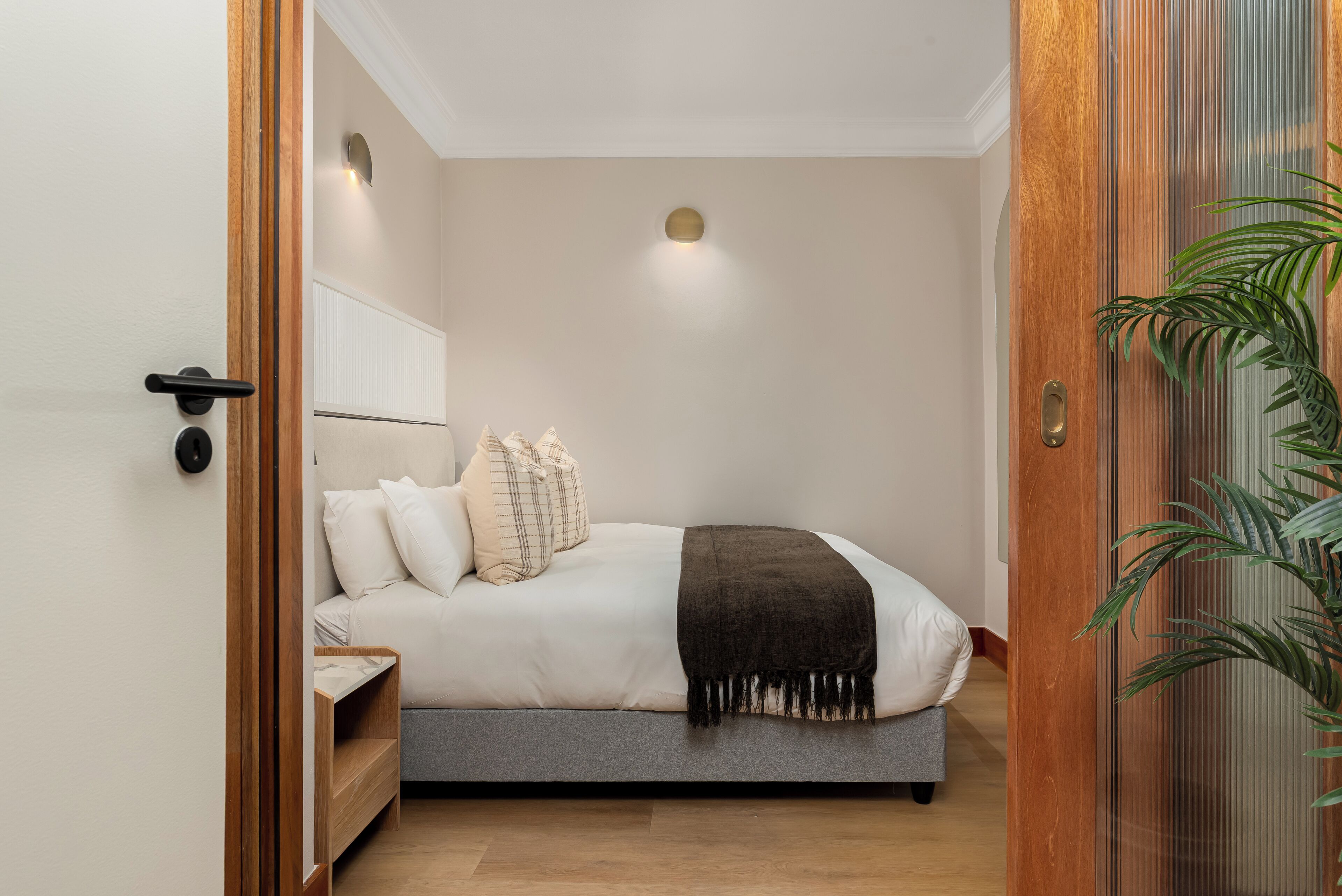 A bedroom with a bed, bedside tables, a nightstand, a wall-mounted light, a door, and a plant.