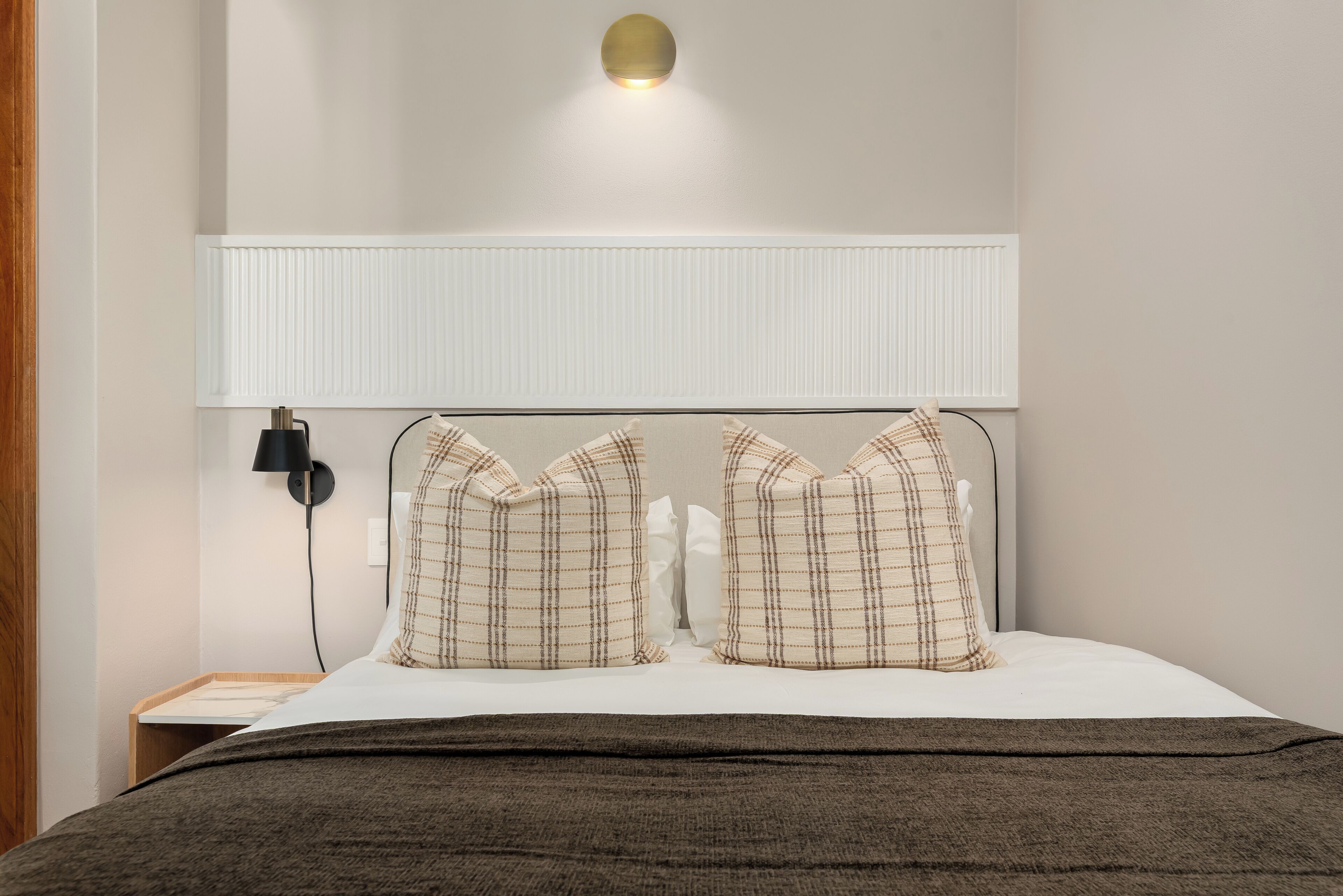 A neatly made bed with a headboard, two pillows, a bedside lamp, and a wall-mounted light fixture.