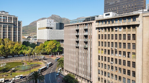 Um edifício alto com vários andares e varandas, rodeado por outros edifícios e uma montanha ao fundo.
